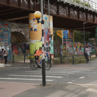 Street under a graffiti-covered railway bridge with cyclists and pedestrians crossing at a zebra crossing.