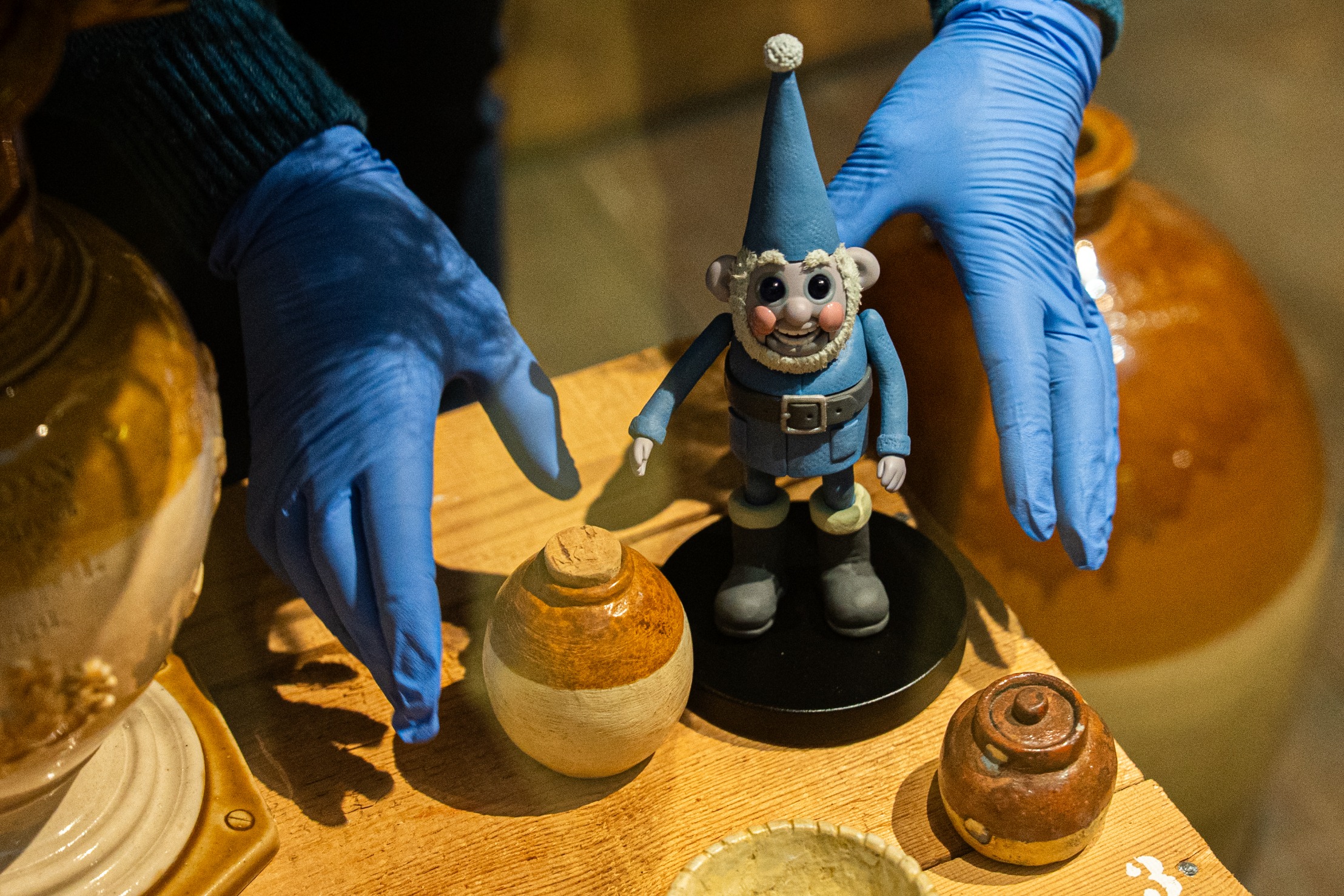 Gloved hands placing a small Aardman-style gnome figurine on a stand beside pottery objects on a wooden table.