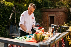 Man in chef whites prepares food outside using healthy ingredients , with a firepit and BBQ to the background alongside greenery and a brick shed.