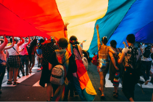 A group of people on a brightly coloured parade holding up a rainbow parachute above their heads