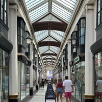 The Arcade in Broadmead, Bristol, with glass-roofed architecture, shopfronts lining both sides, a row of advertising boards down the centre, and several pedestrians walking through the passage.