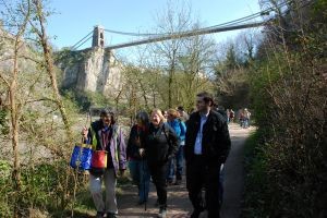 A group of people walking along a wooded riverside path beneath the Clifton Suspension Bridge in Bristol on a sunny day.