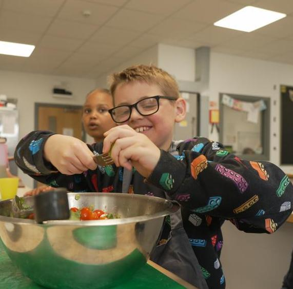 In a kitchen area, a child is wearing glasses and a black jumper with a colourful lego pattern and is holding a fork to squeeze juice into a metal bowl. Other children and adults to the background.