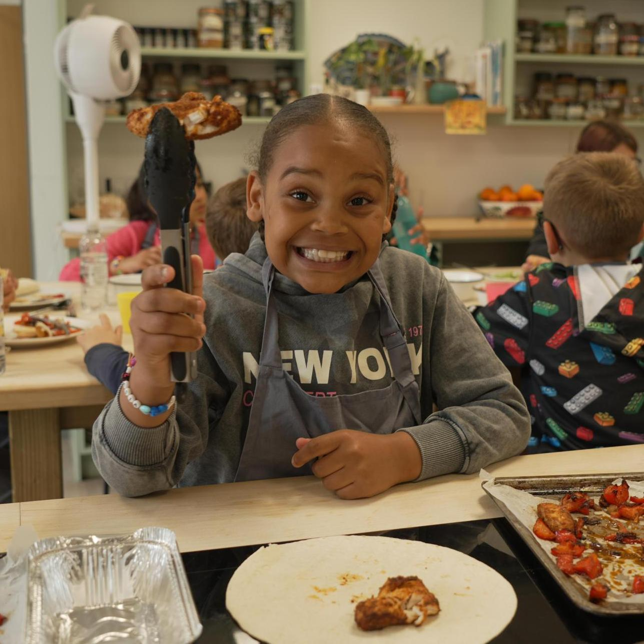 A child in a kitchen is holding food with tongs, looking straight ahead and smiling. Other people in the cooking session are in the background.