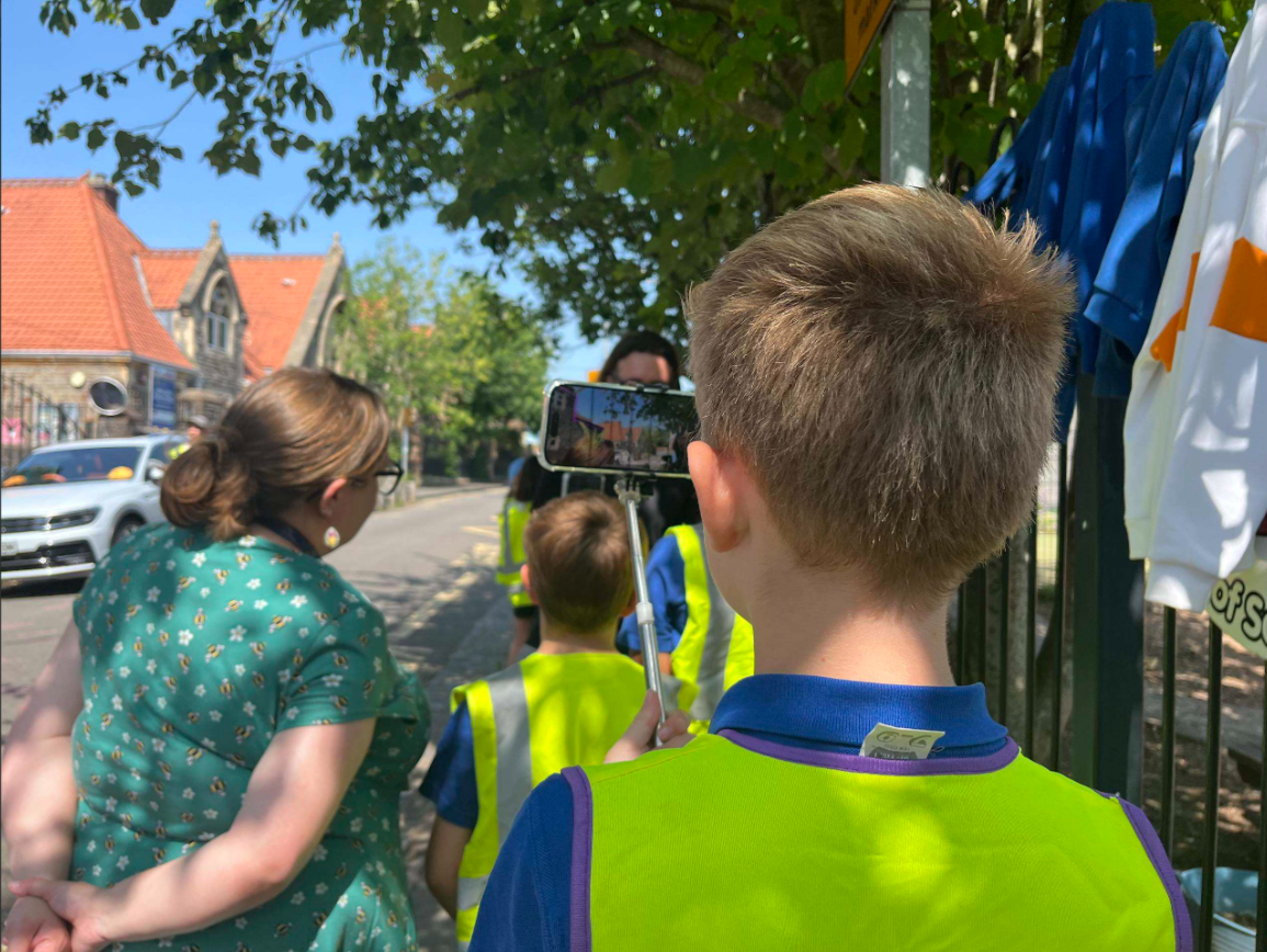 A group of school children wearing high-visibility vests stand on a pavement with an adult while using a smartphone mounted on an extended handheld mount to view street design ideas in augmented reality. Trees provide shade along the road, and school clothing can be seen hanging on a fence beside them, with buildings visible in the background.
