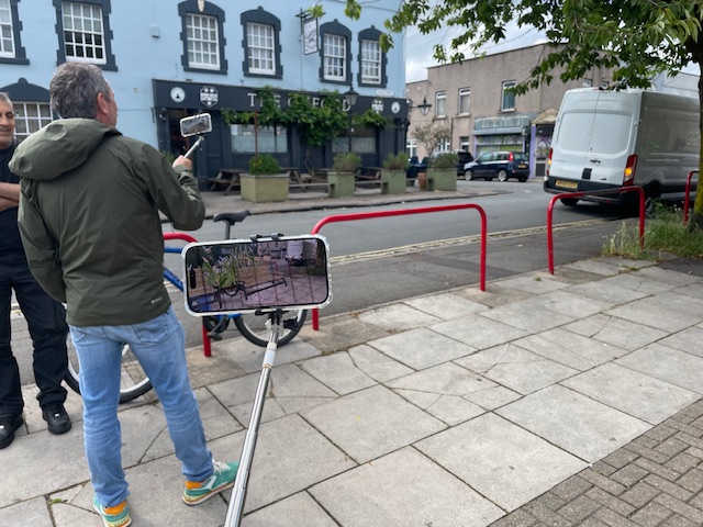 A person uses a smartphone on an extended handheld mount to view proposed street design features, illustrated in augmented reality from a pavement. Red barriers, a bicycle, a parked van and buildings along the street are visible in the background.
