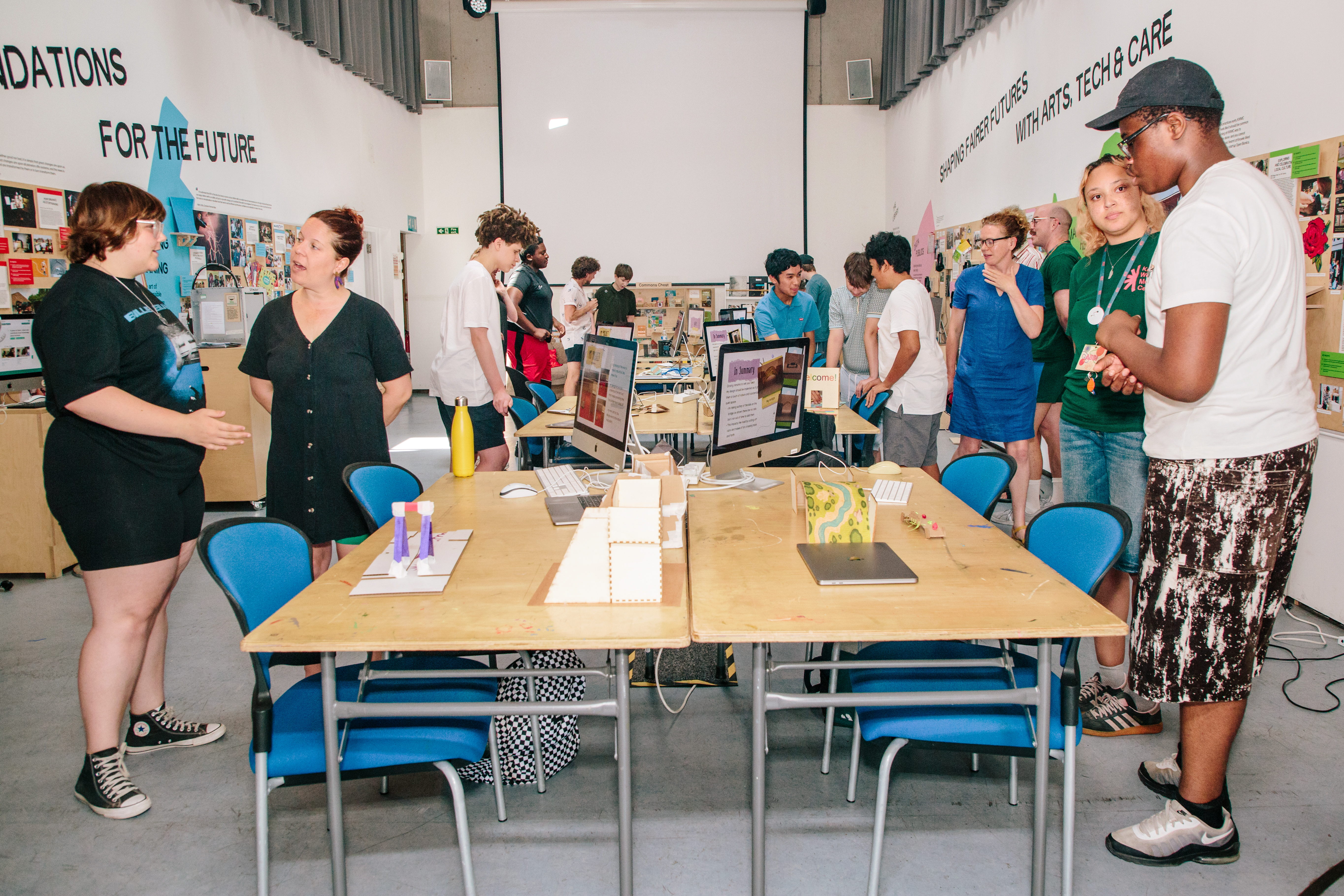 A young person presents a small-scale street design model to Counsellor Ed Plowden during the showcase event of their work experience week focusing on Liveable neighbourhood design. The model, made from cardboard and wooden materials, sits on a table in a meeting room used for discussion and planning activities.