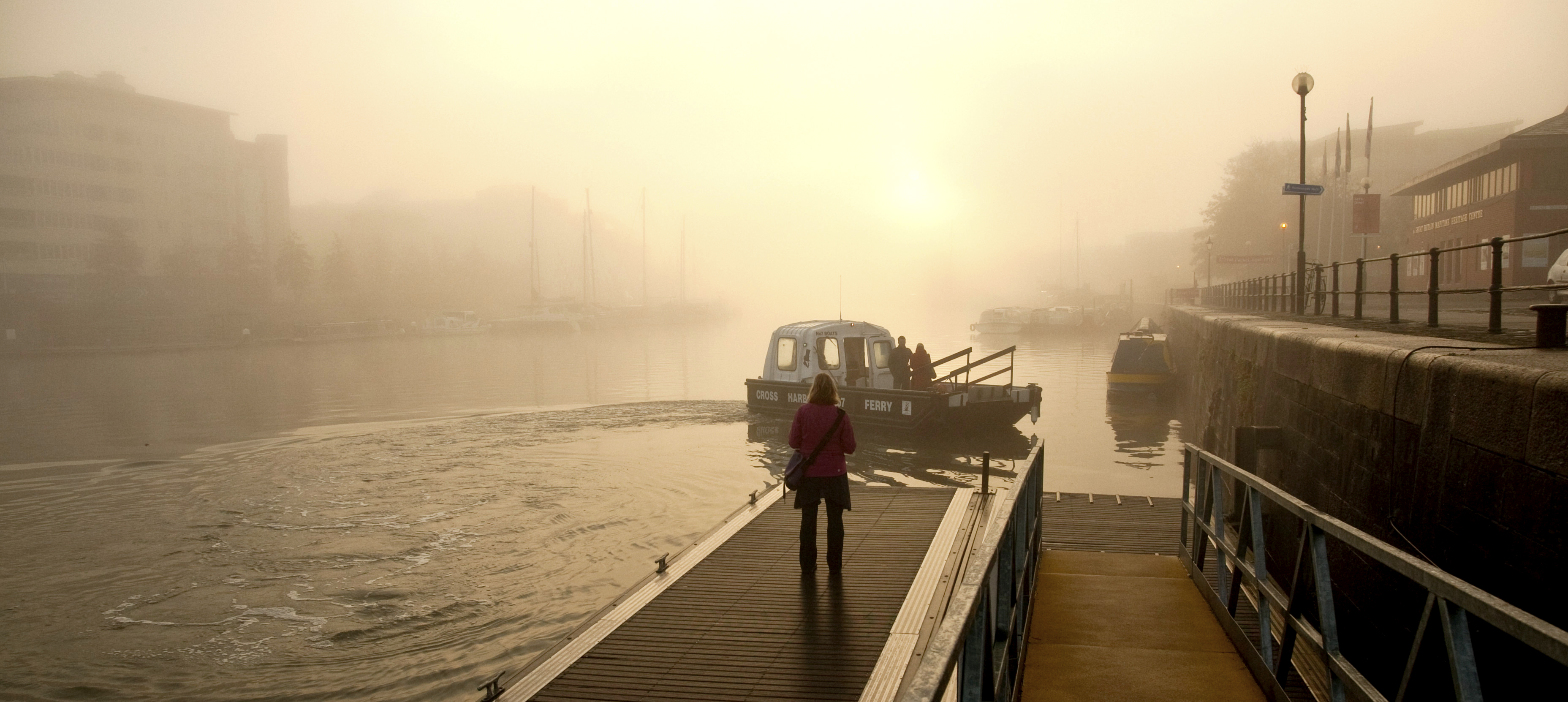 A misty day, a small boat about to cross the water and a lady stood watching the boat