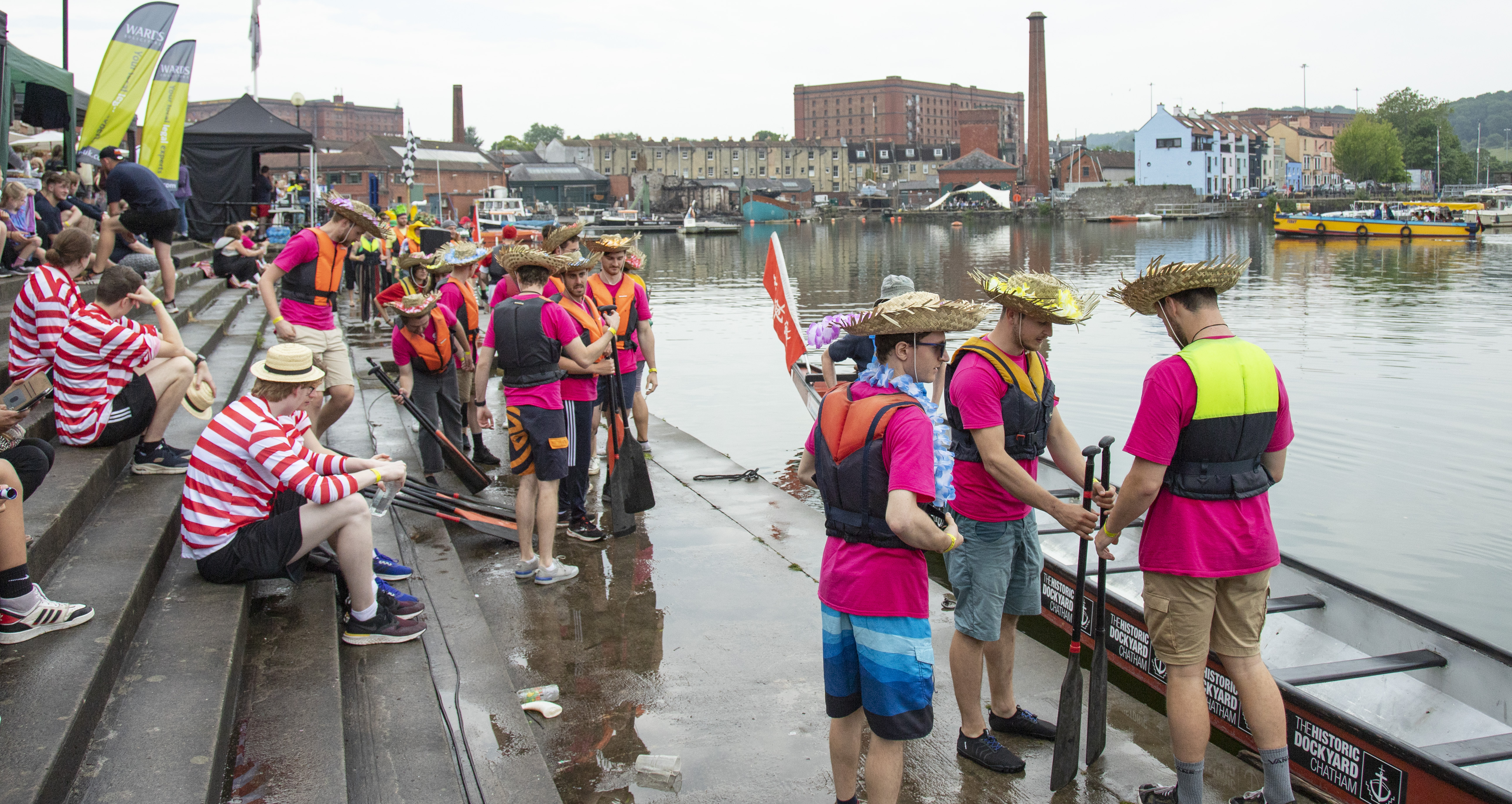 Some people in pink T shirts and other people in red and white striped T Shirts on the shore of the harbour