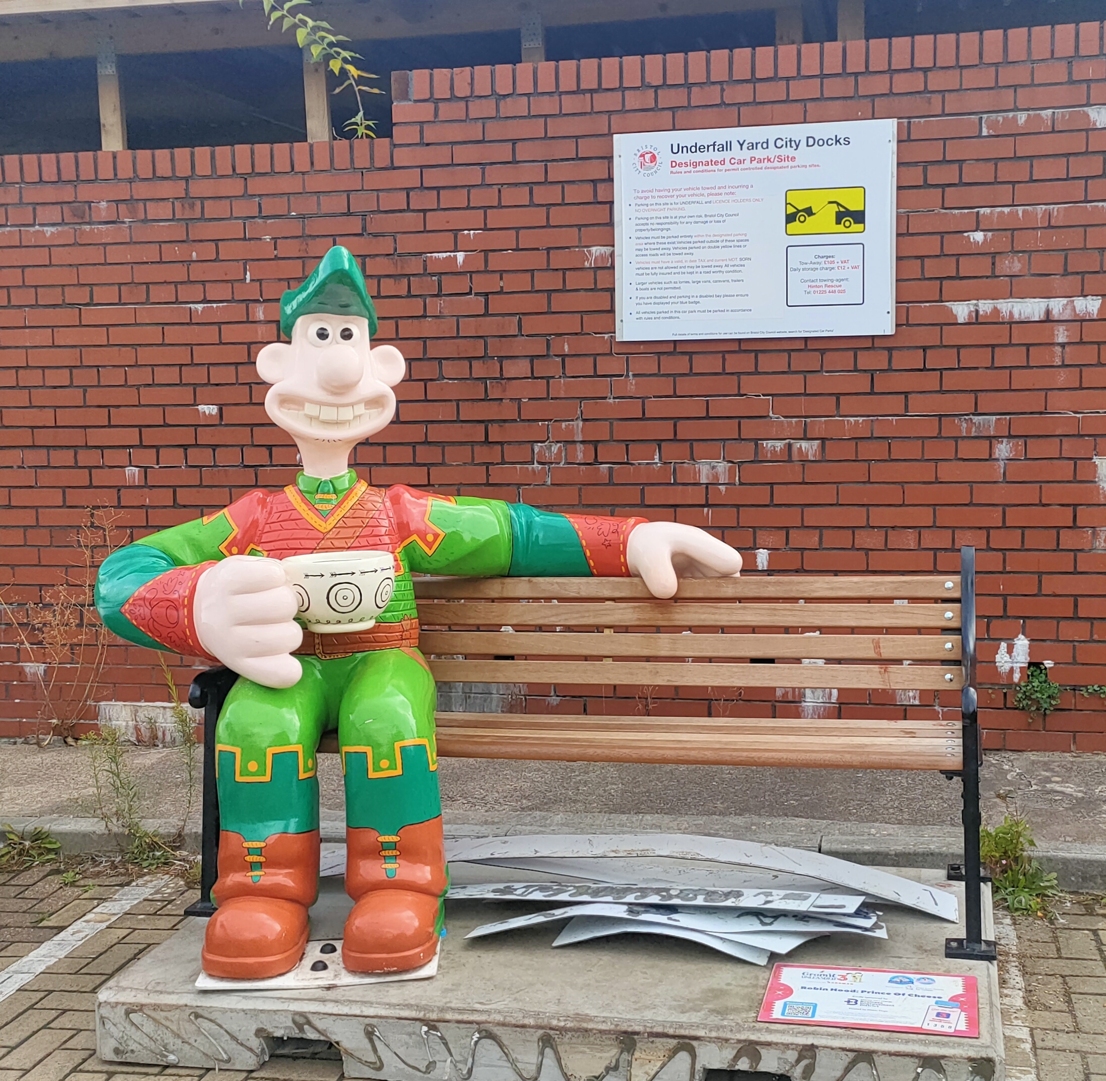 A model of a man in an orange and green outfit. with a cup of tea, sat on a bench