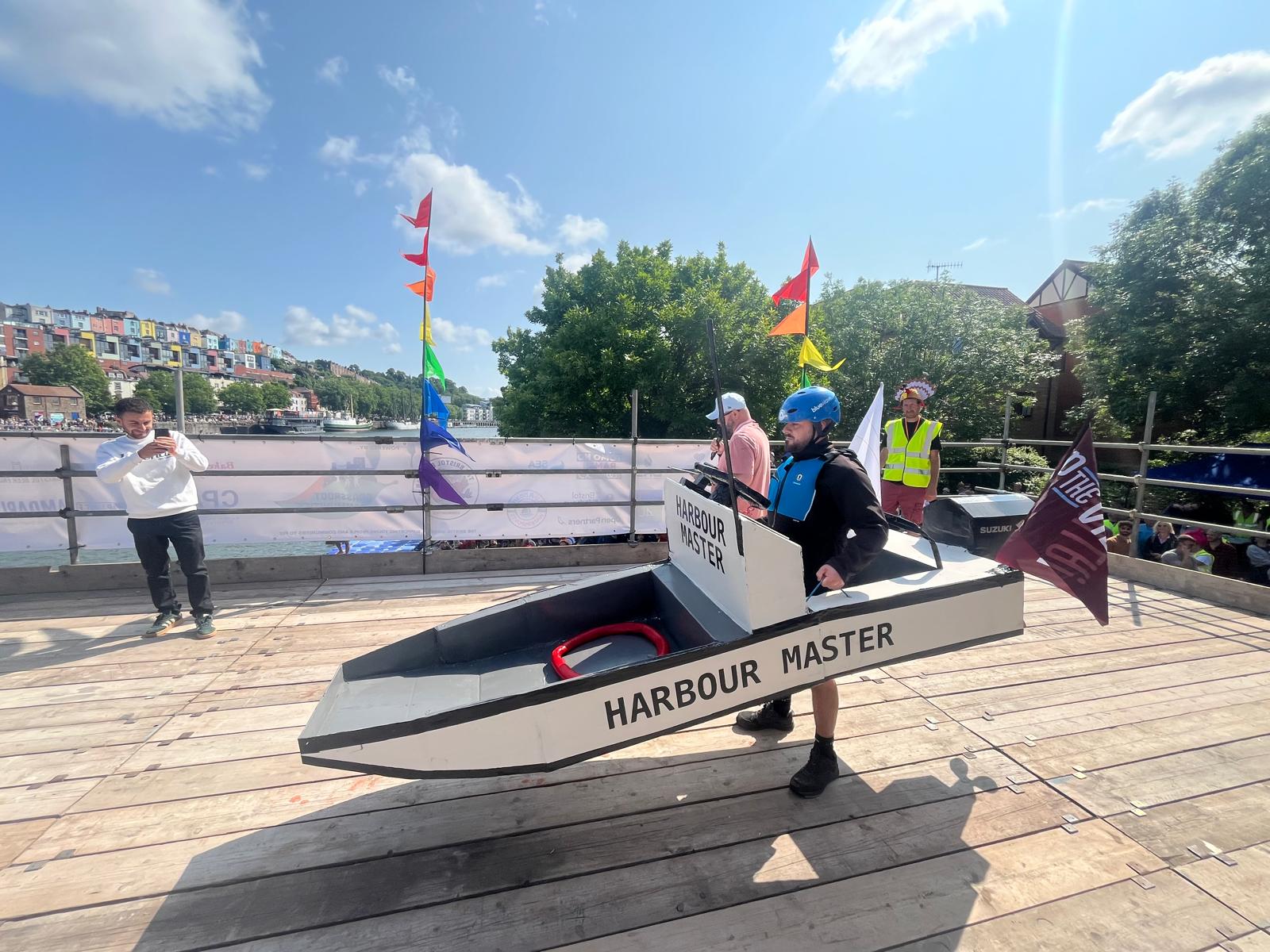 A man in a cardboard boat with wings stood on a platform. Some people in the background, blue skies and clouds
