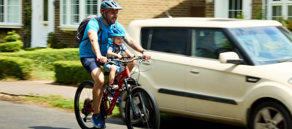 A man on a bike with a child at the front. A cream coloured car in the background