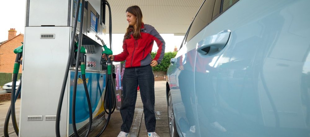 A lady with long brown hair and a red coat filling up petrol in a blue car
