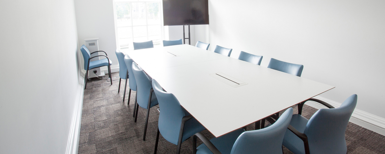 a narrow meeting room with a large white table and blue chairs and a screen in the far end