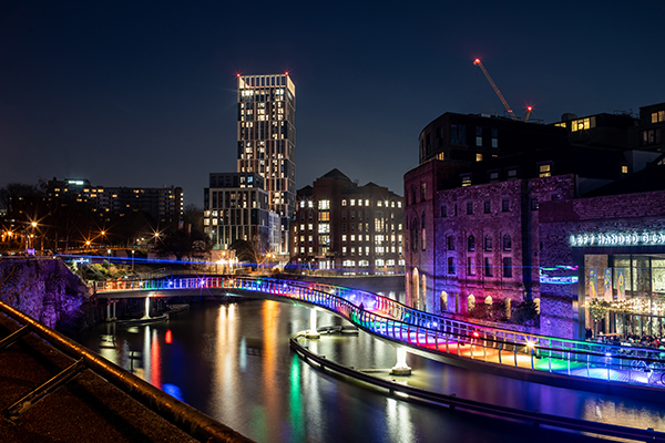 A view from Castle Park Bristol at night showing the multi-coloured lights on the "snake" bridge over the river