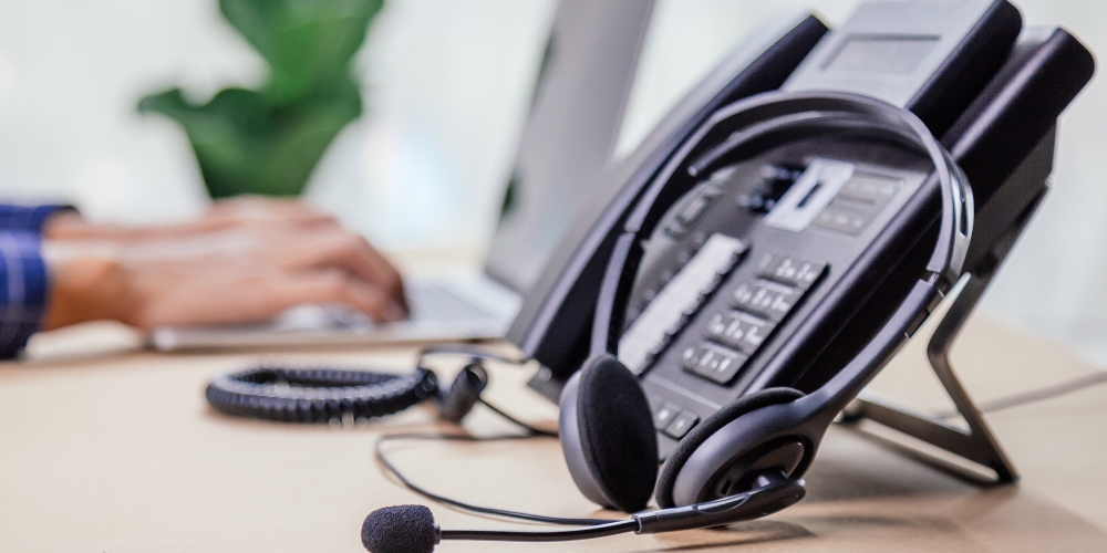 Phone and headset on a desk with a person typing on a laptop in the background