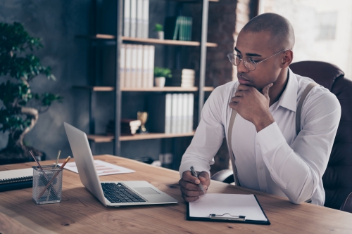 Man sitting at a desk with a clipboard on the desk looking at his laptop 