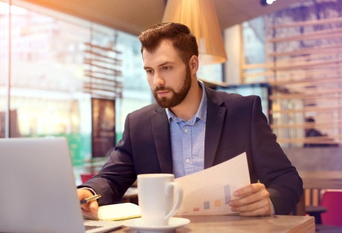 Man sitting at a table in a cafe holding paperwork while looking at a laptop
