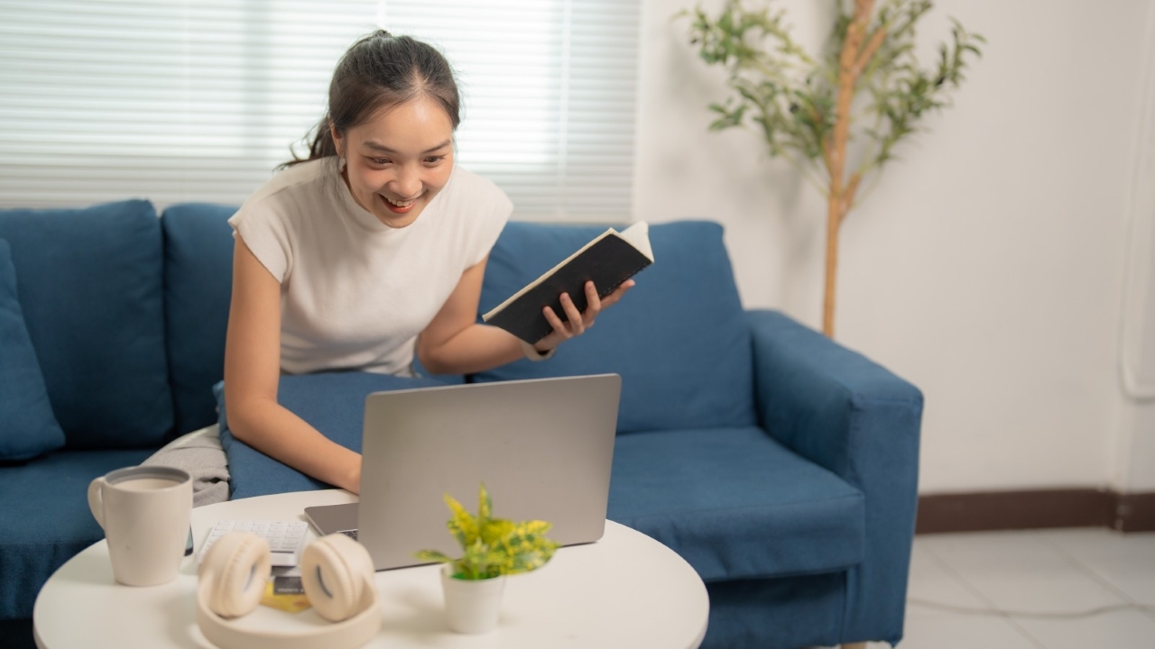 Young woman sitting on a sofa holding a notebook and looking at a laptop
