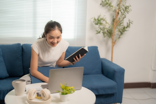Young woman sitting on a sofa looking at her laptop with a notebook in hand