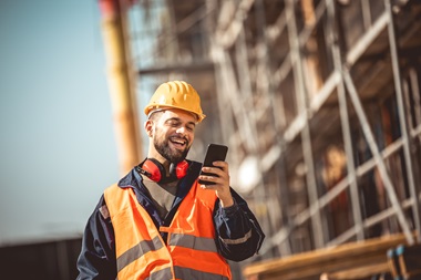 A worker in a hard hat and high vis jacket smiling while looking at his phone.