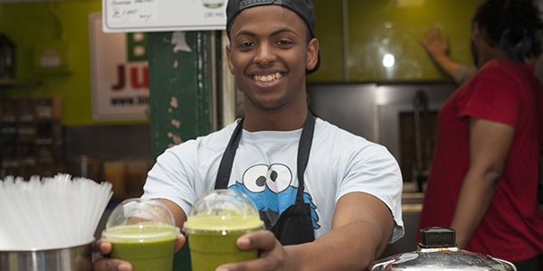 A man smiling, wearing a cookie monster t-shirt and holding out two plastic cups of green juice