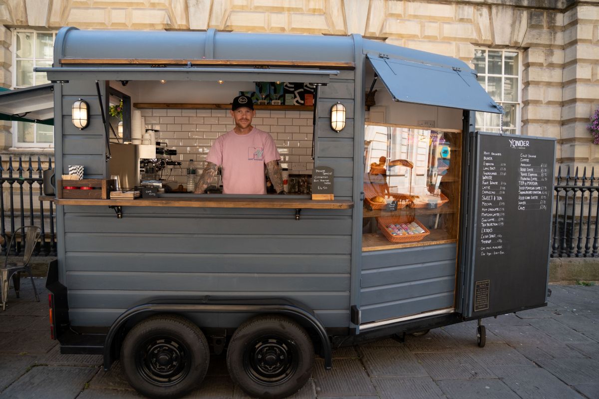 A van with an open hatchett. Inside is a coffee machine and a man