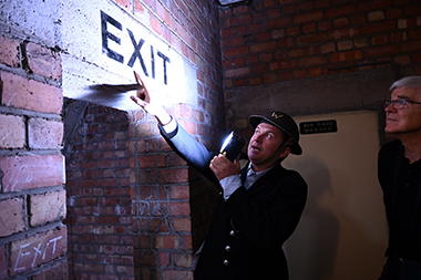 A man dressed as a world war 2 warden shows another man an underground bunker