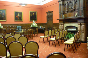 A room with wood panelling, period paintings, a large fireplace and two sets of three rows of chairs