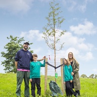 a family with two smiling children stand beside a small tree